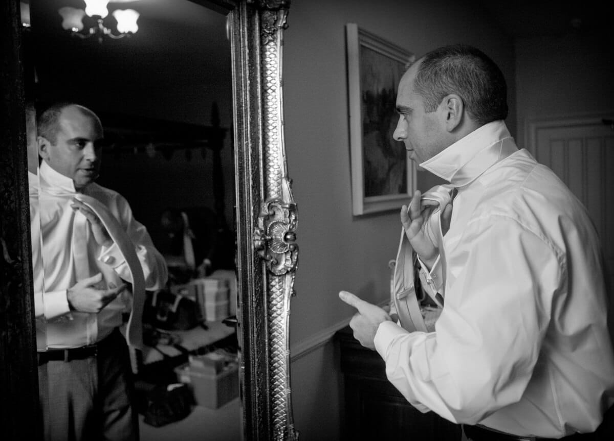 Groom looking in mirror putting on tie at Lemore Manor
