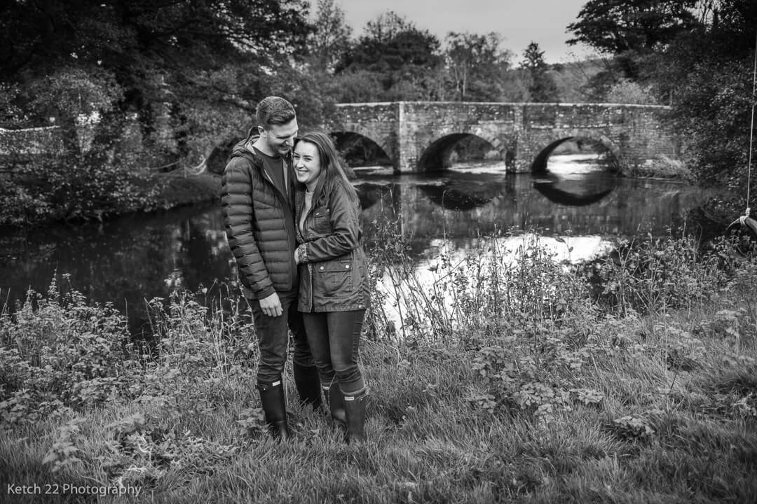 Engaged couple hugging in front of bridge over a river