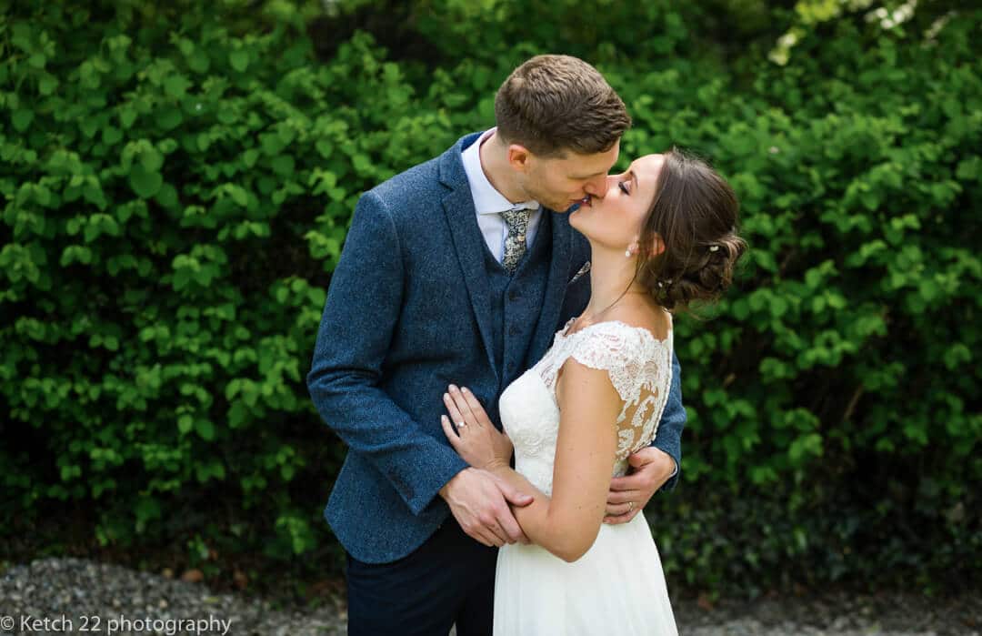 Portrait of bride and groom kissing in front of green bush