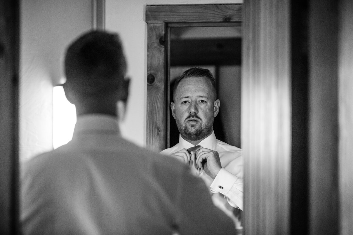 Groom looking in mirror and putting on tie at Cotswold Wedding at Cripps Barn