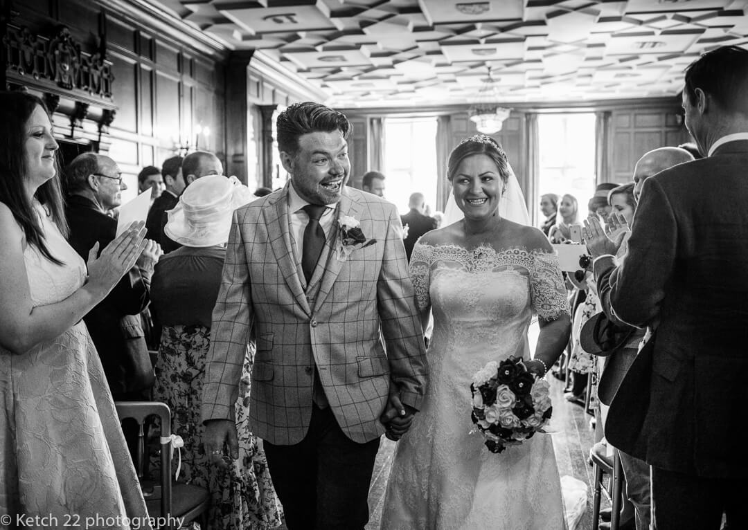 Bride and groom leaving ceremony room with wedding guests cheering