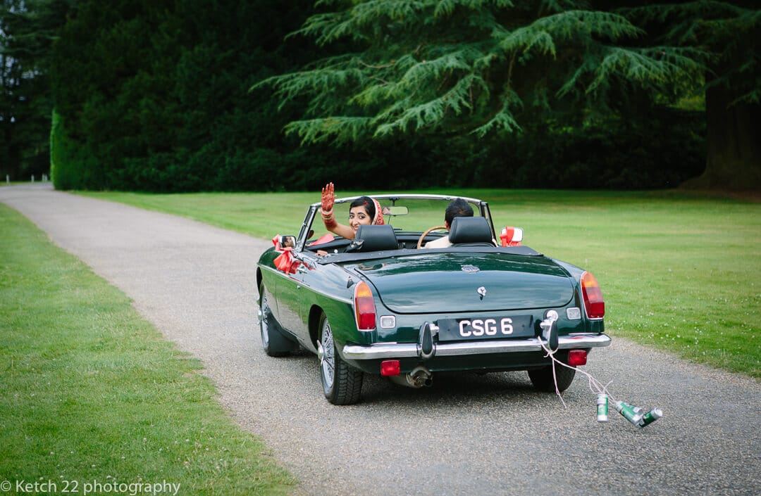 Hindu bride waving from green wedding car with cans trailing