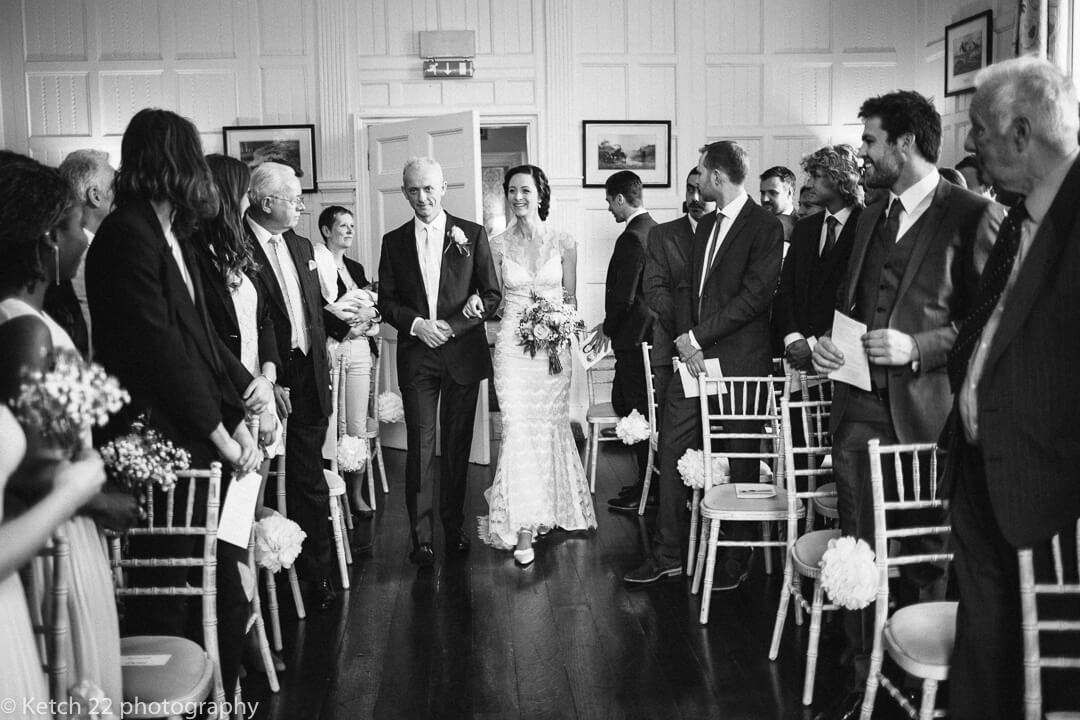 Father and bride enter wedding ceremony room with wedding guests looking on