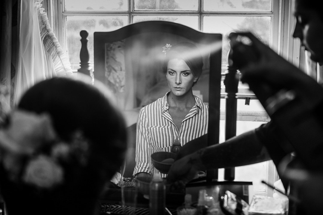 Story telling wedding photograph of bride having her hair sprayed in mirror at bridal preparations