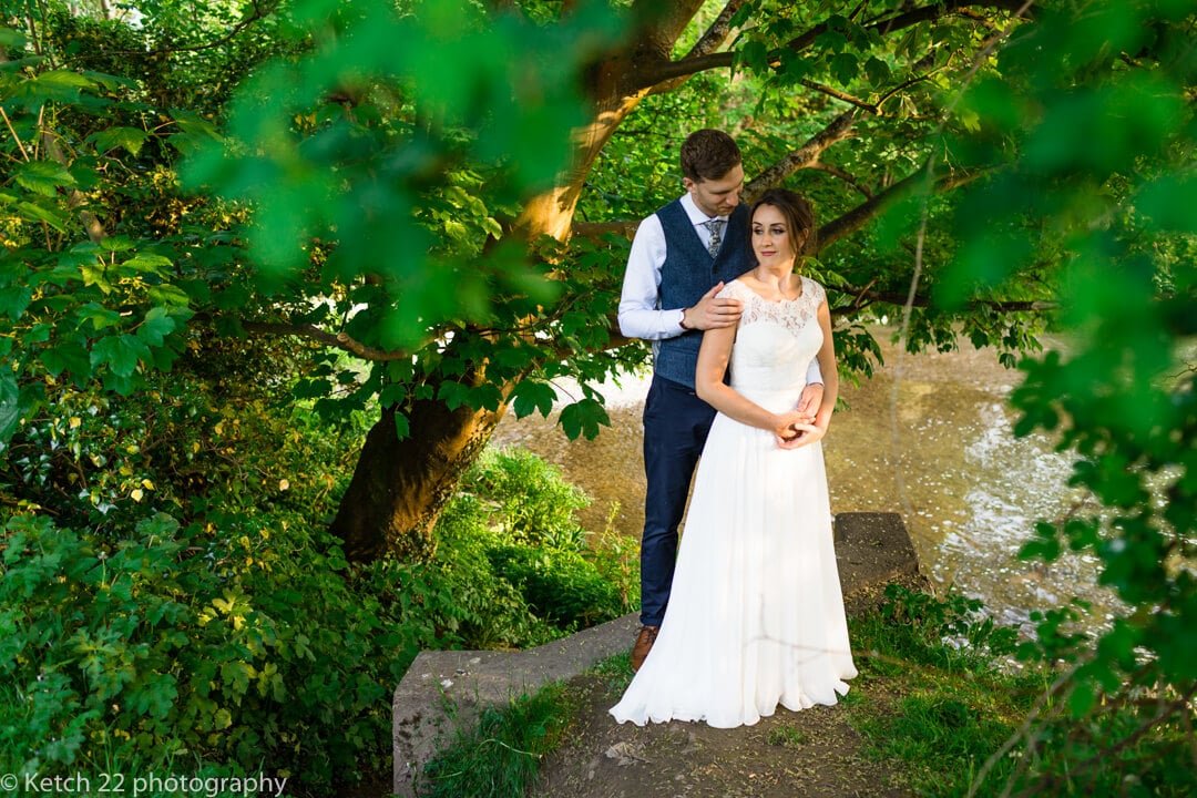 Portrait of bride and groom stood by a river and green trees